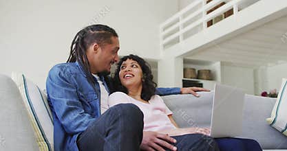 Happy biracial couple sitting on couch using laptop in living room