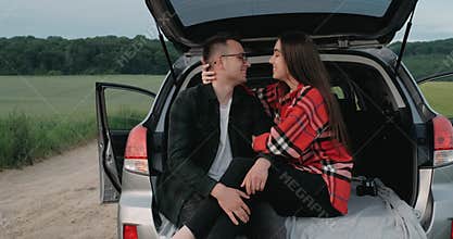 Young Couple, Guy with Girlfriend Sitting in the Opened Car Trunk, Hugging and Smiling while Enjoying Road Trip Through