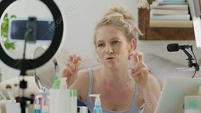 Smiling woman influencer blogger recording a video tutorial on smartphone camera explains what to do to apply makeup cosmetics.
