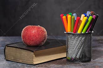 Back to school. Apple in drops of water lying on the book, next to multicolored markers standing in the stand