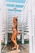 A woman is standing in a shower stall, washing her hair. The shower stall is white and has a wooden frame. The woman is