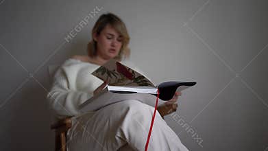 A medium-sized woman in light clothes reads a book while sitting in a white chair in a light room