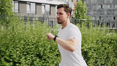 A young concentrated sportive man does morning jog on the sidewalk and checks the time on the watch. An athlete guy