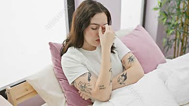 Exhausted young female in pyjamas rubbing her aching eyes, stressed with headache indoors, seated on bed in home apartment bedroom