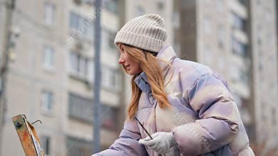 Closeup view of female pretty artist drawing standing on the street using crayon and easel