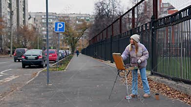 Woman is painting outdoors with a crayon on a wooden easel standing in the city street