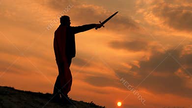 Free male knight. Superman with a sword in his hand and in red cloak stands on a mountain in sunset light. free man