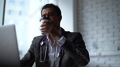 Serious young male doctor working on laptop computer and have break to drink coffee from cup.