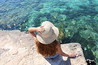 Summer lifestyle image of young woman sitting on the cliff above the sea. Enjoying life and looking at the sea. Crystalline