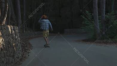 A young guy learns to ride a longboard. It is located in the forest. Dressed in a plaid shirt and a felt hat. Active