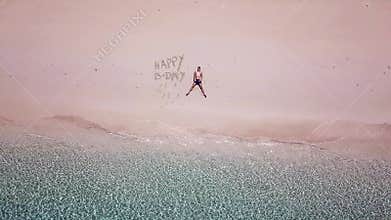 A drone shot of a man lying on a pink sand beach next to `Happy Birthday` sign on a small island near Maumere, Indonesia. Happy