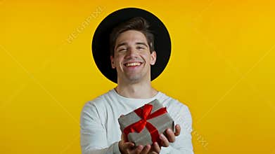 Handsome hipster man holding christmas gift and gives it by hands to camera. He is happy, smiling. Guy in hat on yellow