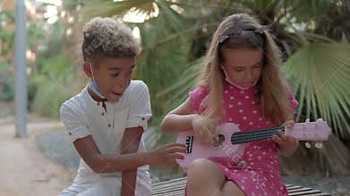 Little caucasian girl plays ukulele sit outdoor with african ethnicity best friend. Adorable pretty multi racial children 10s