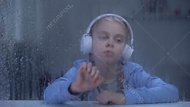 Bored girl in headphones listening to music behind rainy window, home alone