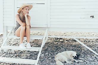 Romantic beautiful woman sits on the steps in front of a white beach house, the dog lies on pebbles under the house.