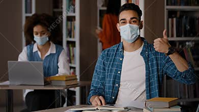 Students in library young indian guy in protective mask sits at desk doing homework preparing for exam writing notes