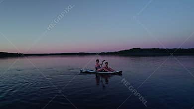 Flying low angle around woman and kids on sup board on calm lake in evening twilight.