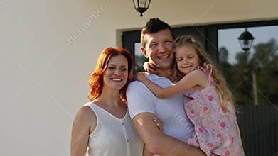 Friendly american dream family demonstrate positive mood outdoors near their house. Father holds daughter in arms and
