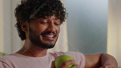 Indian man using laptop when tries green apple. Young guy talking on online meeting and having a lunch. Eating fruits