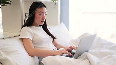 Asian woman working on laptop in bright cozy bedroom setting
