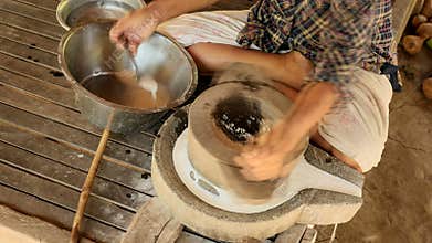 Woman sat cross-legged using hand-turned millstone to grind wet rice for making soaked rice flour 