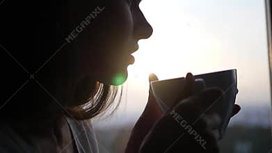 Young attractive girl drinks hot tea or coffee sitting on a window in the rays of a beautiful sunset, with the effect of