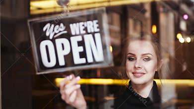 Attractive young blonde woman owner turns sign from close to open and shares a friendly, bright smile to camera. Inside