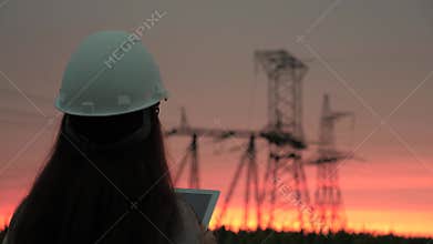 A woman power engineer in white helmet inspects power line using data from electrical sensors on a tablet. High voltage