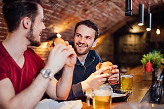 Two happy male friends eating tasty burgers in bar.