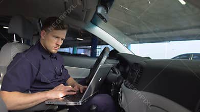 Male police officer working on laptop in car, filling data in crime report