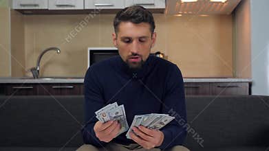 Young man in dark blue pullover and gray pants is sitting on the couch and counting a pack of money. Huge wealth of money