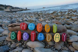 Happy Birthday text composed with multi colored stone letters on wood over a rocks beach