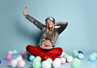 Teen girl in reindeer pajamas, red socks, sleep mask. Yawning, closed eyes, sitting on floor, balloons, blue background. Close up