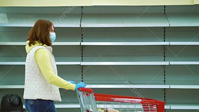 Woman in medical mask walking with shopping cart along empty shelves in store