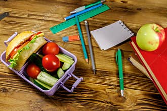 Back to school concept. School supplies, books, apple and lunch box with burgers and fresh vegetables on wooden table
