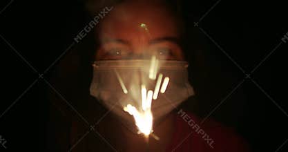 Woman wearing medical face mask holding a lit sparkler with sparks flying. Celebrating fourth, 4th of July during Corona virus