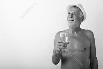 Studio shot of happy senior bearded man smiling and thinking while holding glass of water with hat shirtless against