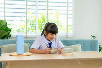 Asian elementary school student girl in uniform eating breakfast cereals with milk and looking cartoon in smartphone to