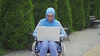 Portrait young muslim woman disabled in a traditional scarf uses a laptop sitting in a wheelchair