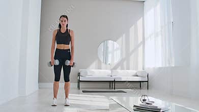 A young athletic woman in a bright apartment performs a deadlift with dumbbells at home, making leans forward to