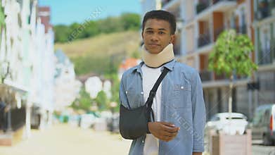 Sad mixed-race young man in foam cervical collar and arm sling sighing on camera