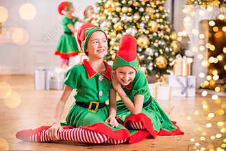 Two cheerful happy girls-friends in costumes of Santa`s elves on the background of a Christmas tree and other children