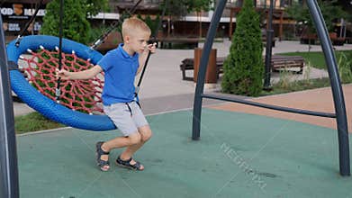 The boy swings on a round wicker swing in the yard on the playground.