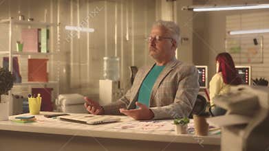 Aged man relaxes in a zen pose and performs the calming breathing practice. A businessman in a stylish jacket works in