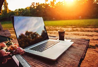 Laptop computer on rough wooden table with coffee cup and bouquet of peonies flowers in outdoor park