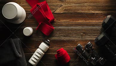 Grey towel Protein shake bottle Dumbbell plate White jar with whey protein Red hand wrap Sneakers on wooden floor