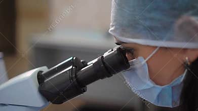 Close up of female scientist in medical protective mask and cap working in research laboratory using microscope. Woman