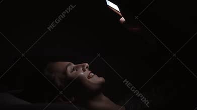 Young smiling woman laying on a couch and using her phone in a dark room, chatting in the darkness with smartphone