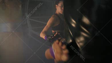 Caucasian strong female kickboxer exercising with a boxing bag in dark gym with smoke and kicking a boxing bag with her