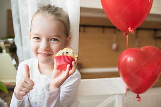 Cute preschooler girl celebrating 6th birthday. Girl eating her birthday cupcake in the kitchen, surrounded by balloons.